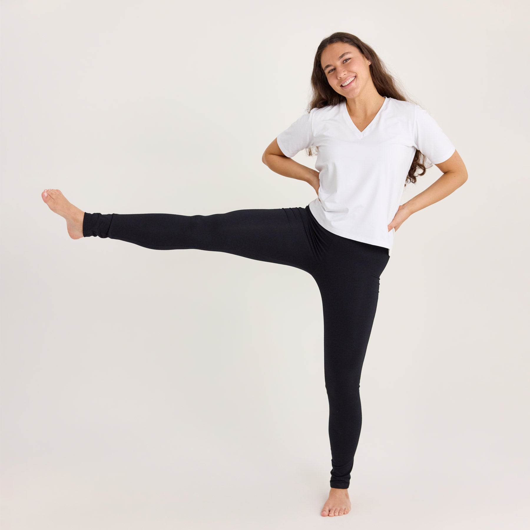 Mallilla on mustat bambuleggingsit ja valkoinen Jokapaikan t-paita. Malli nostaa toista jalkaansa ylös korostaen leggingsien joustavuutta.