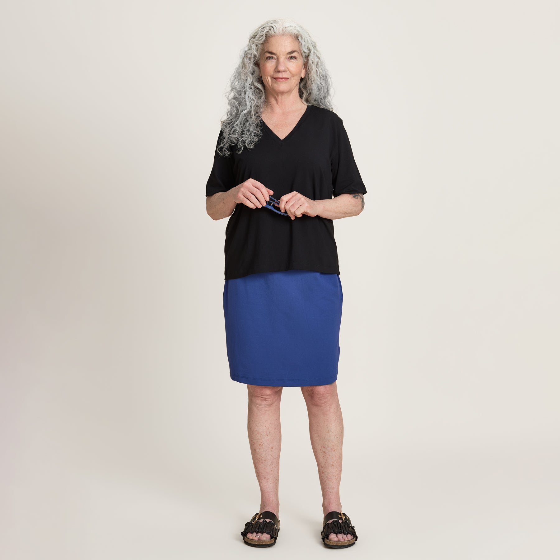 Harmaahiuksinen nainen hymyilee lempeästi mustassa Jokapaikan t-paidassa, ja Jokapaikan hameessa.