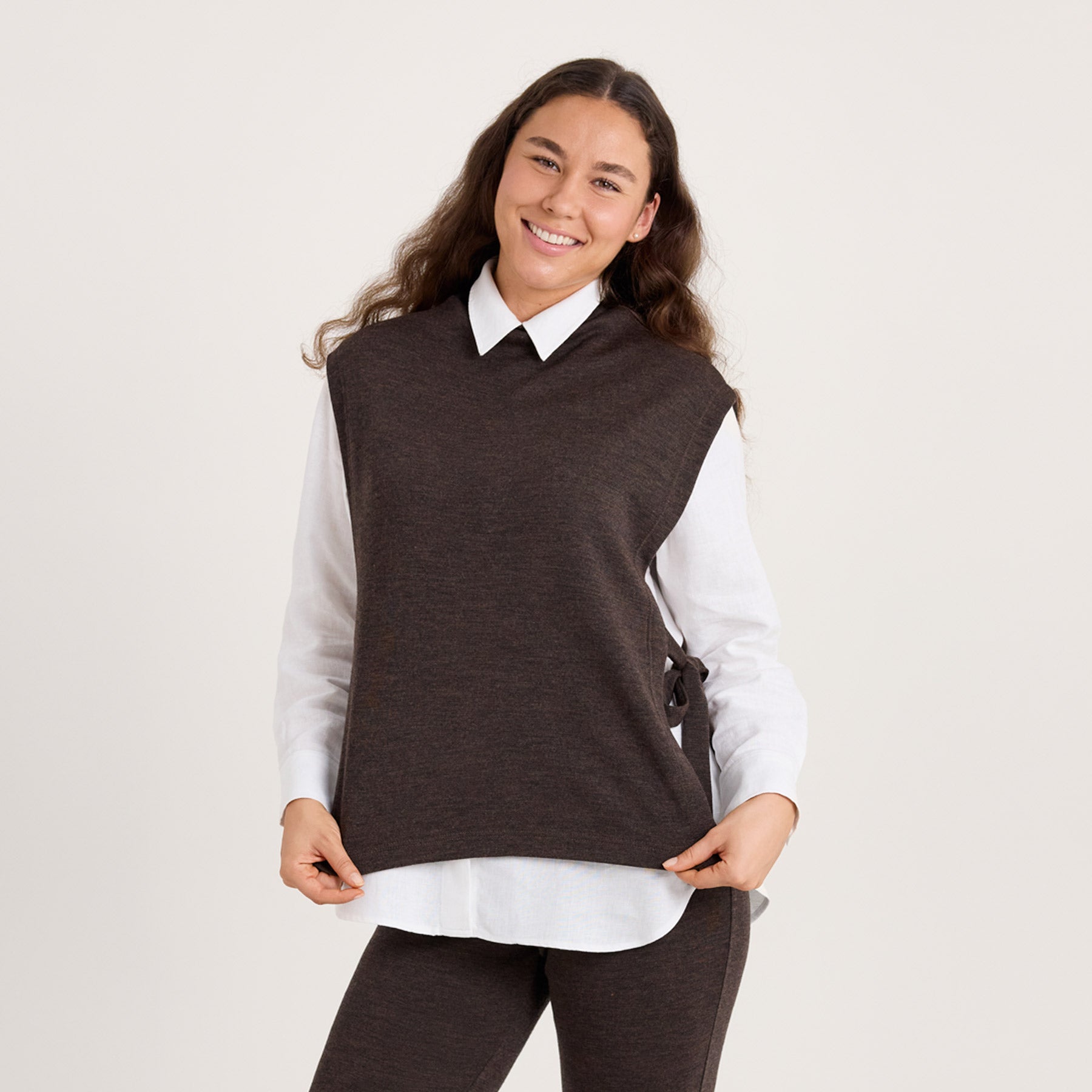 Lähikuva meleeratun ruskean värisestä liivikaulurista. Malli pitää käsillään kiinni helmasta. 100% merinovillaa, joka ei kutita.