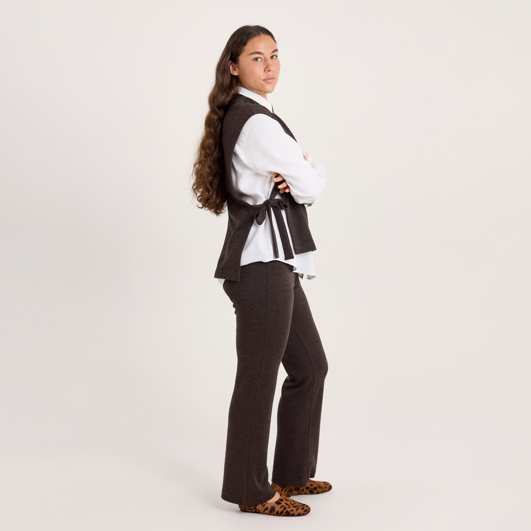 Sivuttain kuvattuna meleeratun ruskean värinen liivikauluri ja rennot neulehousut. Liivikaulurin alla on valkoinen kauluspaita.