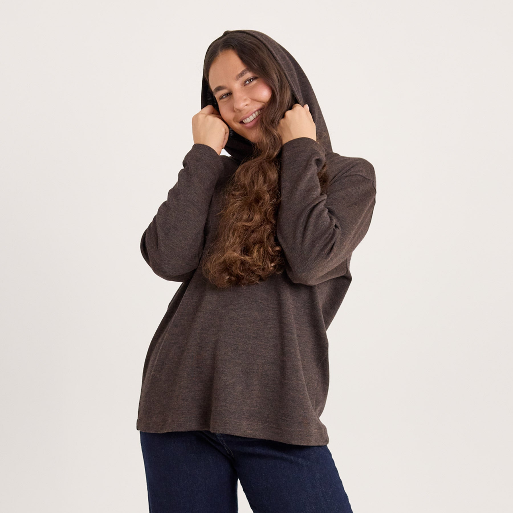 Lähikuva meleeratun ruskeasta suorasta hupparista. Mallilla on huppu päässä, ja hän pitää käsillään hupun reunoista kiinni. Valmistettu Kangasalla.