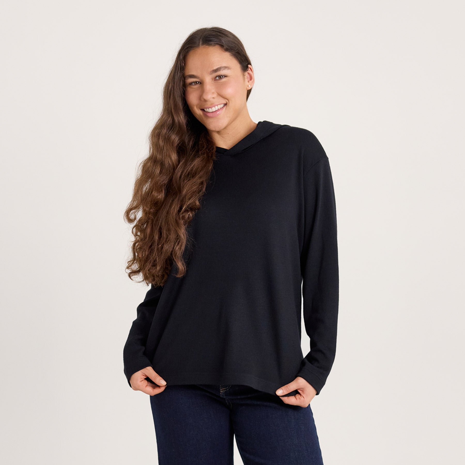 Lähikuvassa mustan värinen suora huppari. Malli pitää käsiä hupparin helmassa, ja tuo esille hupparin suoran linjan.