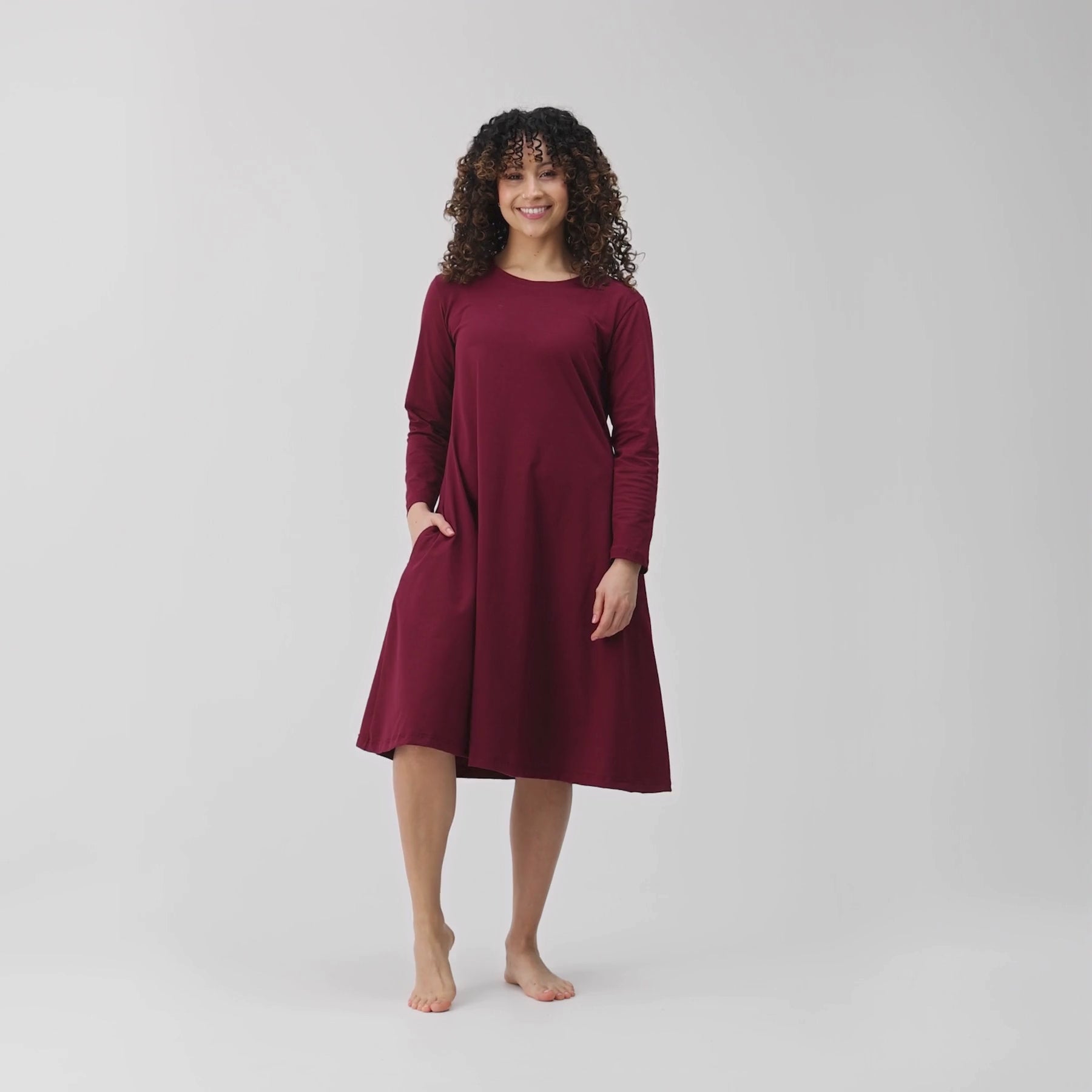 Videolla on viininpunainen kellomekko. Malli esittelee mekon taskun ja hulmuavan kellohelman.