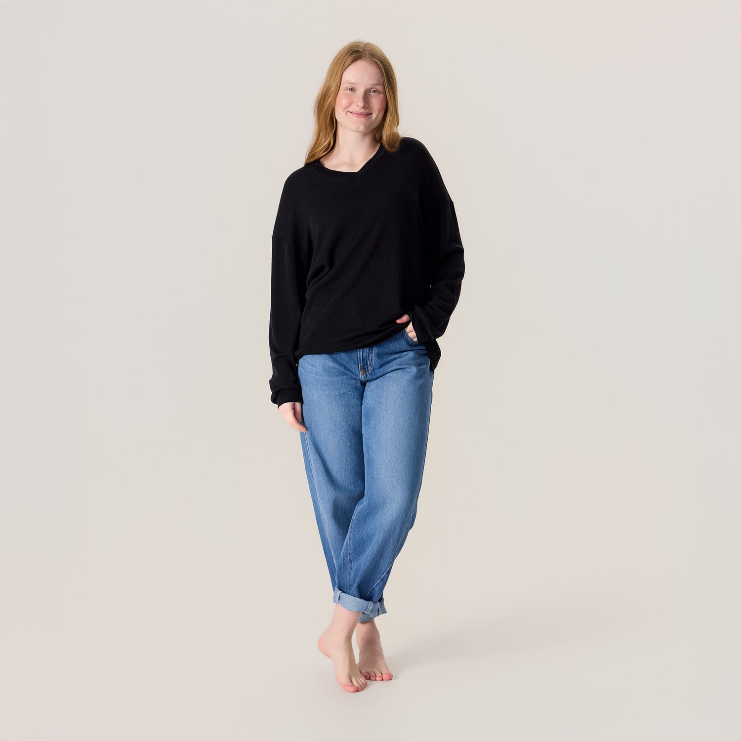 Nainen farkuissa ja unisex-mallisessa mustassa neulepaidassa. Neulepaidassa on v-pääntie ja pitkät hihat.