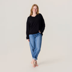 Nainen farkuissa ja unisex-mallisessa mustassa neulepaidassa. Neulepaidassa on v-pääntie ja pitkät hihat.