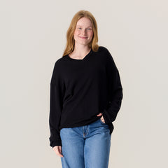 Lähikuvassa nainen farkuissa ja unisex-mallisessa mustassa neulepaidassa. Neulepaidassa on v-pääntie ja pitkät hihat.